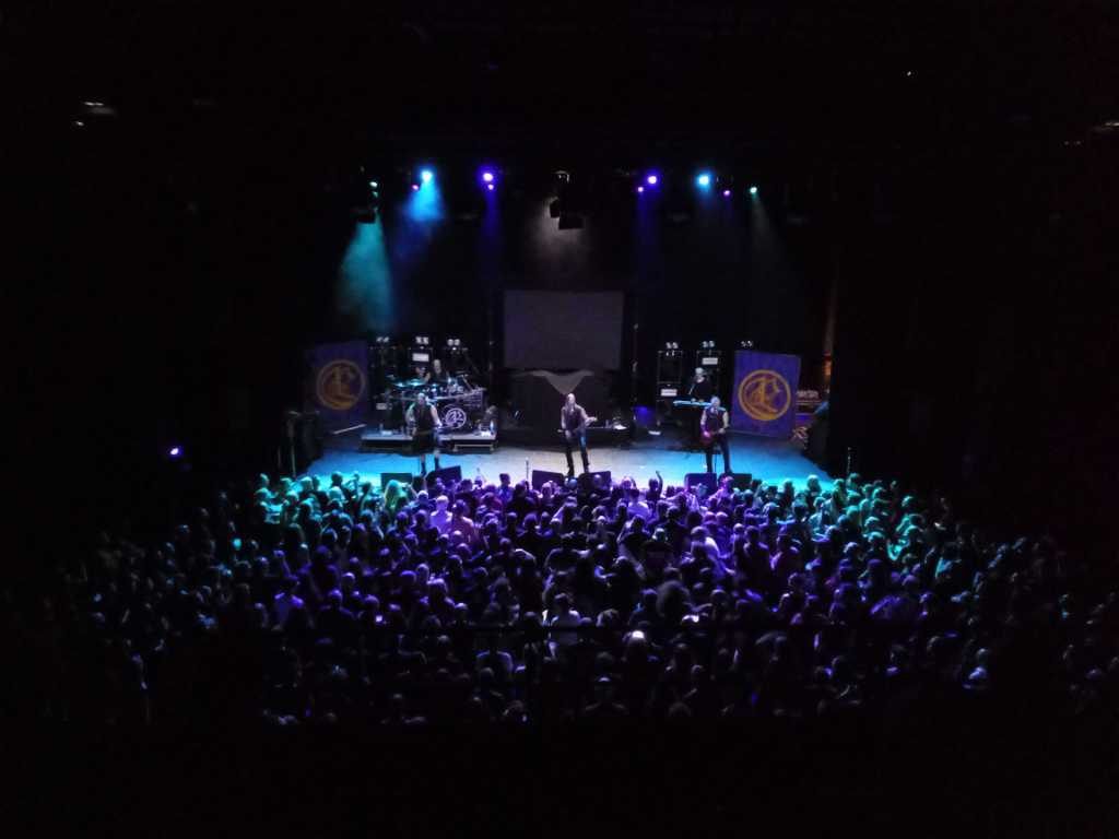 Ensiferum sur scène. Le batteur au fond à gauche. Le bassite à gauche, le chanteur au milieu. Le second guitariste à droite. En guise de décor, deux drapeaux avec le logo du groupe, de part et d'autre de la scène.